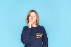 Woman wearing a navy Unique & Loved sweatshirt in front of a bright blue background. She has her finger on her chin and is looking up to her right as if in thought.