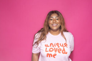 Woman wearing a pink Unique & Loved sweatshirt in front of a bright pink background. She is looking directly at the camera and smiling.