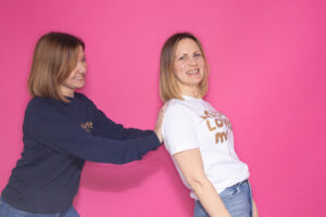 Two women in Unique & Loved tops in front of a bright pink background. Both women are side on to the camera, the woman on the right is leaning backwards and is supported by the hands of the woman on the left.